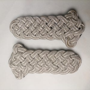 Puede incluir: Un par de hombreras de color plateado con un intrincado diseño de cordón trenzado. Cada pieza tiene un cuerpo rectangular con un extremo más ancho y redondeado, construido con hilo metálico tejido en un patrón denso de bucles entrelazados.