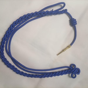 Puede incluir: Un cordón trenzado de color azul brillante que presenta un nudo decorativo en un extremo y una punta cilíndrica de tono dorado metálico. El cordón está hecho de un material sintético liso e incluye un nudo deslizante para ajustar la longitud.