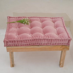 May include: A rectangular, tufted pink velvet floor cushion with a piped edge, resting on a light-colored wooden bench. The cushion features a grid of fabric-covered buttons and is accented with a small sprig of white baby's breath flowers.