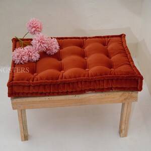 May include: A rectangular, tufted, burnt-orange velvet floor cushion with a thick, piped edge, resting on a light-colored wooden bench. Three artificial pink flower heads are placed on the left side of the cushion. The text LIVINGSTERS is visible.