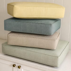 Puede incluir: Una pila de cuatro cojines de asiento rectangulares con un acabado de tela texturizada. Los cojines son de color amarillo pálido, azul apagado, beige claro y verde salvia suave, cada uno con bordes ribeteados y una forma estructurada.