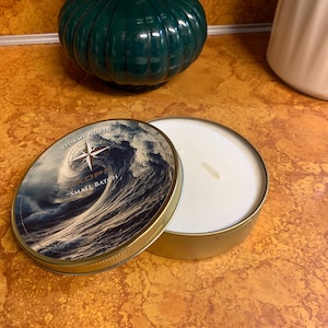 May include: A small batch candle in a gold tin with a lid featuring a wave design and the text "Stormy North". The candle is white, and the lid is open. The candle is on a textured orange surface.