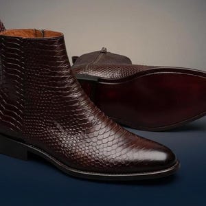 Könnte beinhalten: Ein Paar dunkelbraune Stiefeletten mit einer strukturierten Schlangenledermusterung. Ein Stiefel steht aufrecht und zeigt einen seitlichen Reißverschluss sowie eine Zuglasche an der Ferse, während der andere Stiefel flach liegt und eine glatte Sohle zeigt.
