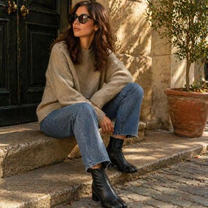 May include: A person wearing a beige sweater, blue jeans, and black sunglasses sits on stone steps. The person is also wearing black leather split-toe ankle boots. A dark wooden door and a potted plant are in the background.