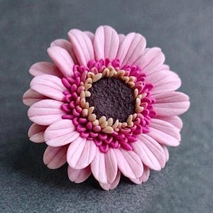 May include: A sculpted flower brooch featuring layered light pink petals, a ring of smaller magenta petals, and a central dark brown disc with beige accents. The floral design is crafted with a smooth, matte finish and detailed, realistic petal textures.