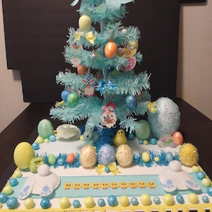 May include: A light blue Easter tree decorated with colorful eggs, bows, and decorative ornaments. The base features a white and blue design with Easter-themed decorations, including bunnies and chicks. The tree is adorned with a large blue bow at the top.