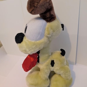 May include: A soft toy dog with a cream-coloured body, a brown head and ears, and a red tongue. The dog has black spots and a white face. The toy is designed to be soft and cuddly, suitable for children or collectors.