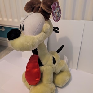 May include: A soft toy dog with a light yellow body, brown ears, and a red tongue. The dog has black spots and a black nose. A tag is attached to the ear. The toy is sitting on a white surface.