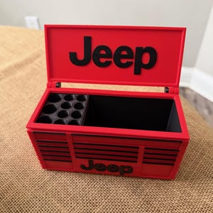 May include: A red Jeep-branded toolbox with a black interior and black accents. The toolbox has a hinged lid with the word "Jeep" in large black letters. The front of the toolbox also features the word "Jeep".