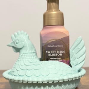 Peut inclure: Un porte-savon en forme de poule bleu clair contenant un flacon de savon moussant pour les mains Sweet Mum Blossom de Bath & Body Works. Le flacon de savon a une pompe dor&eacute;e et une &eacute;tiquette florale. Le tout est sur une surface en bois.