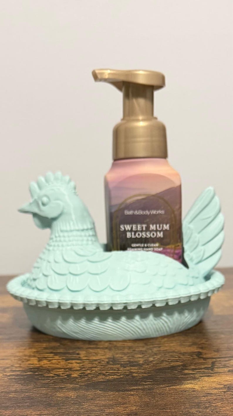 Peut inclure: Un porte-savon en forme de poule bleu clair contenant un flacon de savon moussant pour les mains Sweet Mum Blossom de Bath & Body Works. Le flacon de savon a une pompe dor&eacute;e et une &eacute;tiquette florale. Le tout est sur une surface en bois.