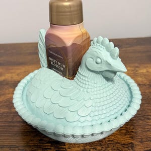 Peut inclure: Un porte-savon en forme de poulet bleu clair avec une bouteille de savon Sweet Mum Blossom. Le support pr&eacute;sente un design d&eacute;taill&eacute; et une pompe dor&eacute;e. La bouteille de savon porte une &eacute;tiquette avec un paysage.