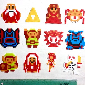 May include: A collection of colorful pixel art characters and symbols, likely from a video game. The figures are made from small, fused beads in various colors, including red, yellow, blue, and white. A black marker and ruler are included for scale.