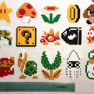 Puede incluir: Una colecci&oacute;n de personajes y objetos coloridos de pixel art de la serie de videojuegos Super Mario. Incluye a Mario, Luigi, una estrella, una seta, una moneda, un bloque de interrogaci&oacute;n y otros elementos ic&oacute;nicos. Se incluye un rotulador negro y una regla.