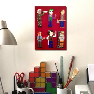 Peut inclure: Une toile rouge avec de l'art pixel de six personnages, dont un zombie et le Joker. L'œuvre est accrochée à un mur blanc au-dessus d'un bureau avec une lampe, des tasses et des blocs géométriques colorés.