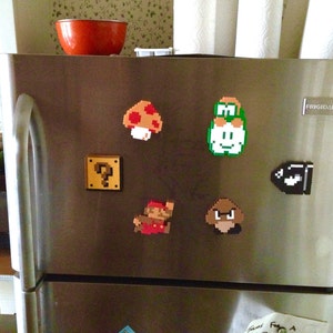 Puede incluir: Puerta de nevera de acero inoxidable adornada con imanes pixelados con personajes e iconos de la serie de videojuegos Super Mario. Incluye un champi&ntilde;&oacute;n, un bloque de interrogaci&oacute;n, Mario, Luigi, un Bill Bala y un Goomba.