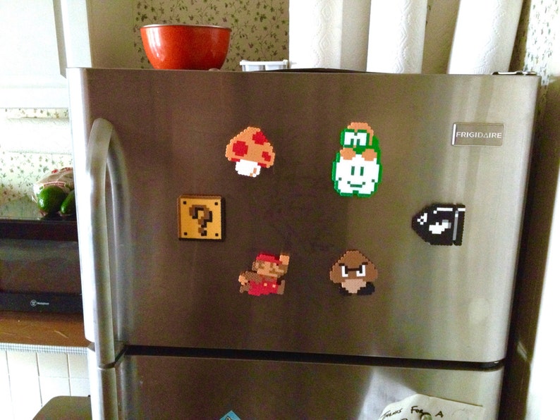 Puede incluir: Puerta de nevera de acero inoxidable adornada con imanes pixelados con personajes e iconos de la serie de videojuegos Super Mario. Incluye un champi&ntilde;&oacute;n, un bloque de interrogaci&oacute;n, Mario, Luigi, un Bill Bala y un Goomba.