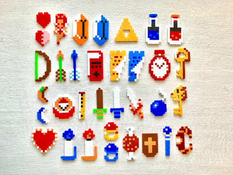 Puede incluir: Una colecci&oacute;n de sprites de pixel art de un videojuego, que incluye corazones, un escudo, un arco y flechas, espadas, llaves y pociones. Los sprites est&aacute;n hechos de cuentas de colores y dispuestos sobre una superficie clara.
