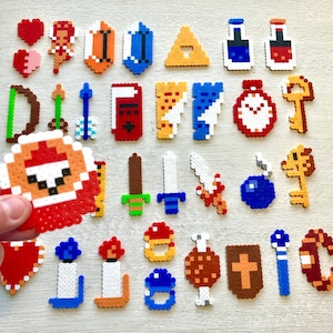 May include: A collection of colorful pixel art designs, including hearts, shields, swords, keys, and other game-themed elements. The designs are made from small, colorful beads, with a hand holding one of the pixel art designs.