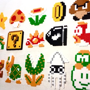 Puede incluir: Una colecci&oacute;n de personajes y objetos coloridos de pixel art de la serie de videojuegos Super Mario. Incluye una estrella amarilla, una seta, una moneda, un Goomba, un Bill Bala y personajes de Mario. El arte est&aacute; hecho de peque&ntilde;as cuentas de pl&aacute;stico fusionadas.