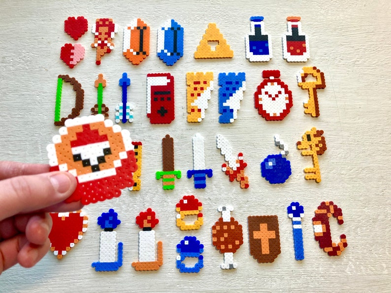 Puede incluir: Una colecci&oacute;n de dise&ntilde;os de pixel art, que incluyen corazones, un hada, un arco y flechas, espadas, llaves y otros elementos tem&aacute;ticos de juegos. Los elementos est&aacute;n hechos de cuentas de pl&aacute;stico de colores, con una mano sosteniendo uno de los dise&ntilde;os.
