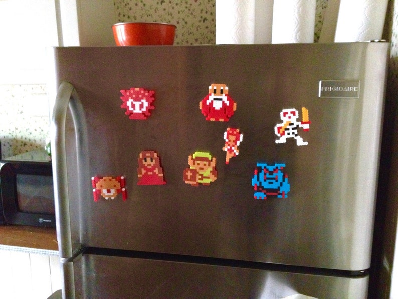 May include: A collection of pixel art magnets in various colors, including red, yellow, blue, and white, are displayed on a stainless steel refrigerator. The magnets depict characters from a video game, including a skeleton, a wizard, and a princess.