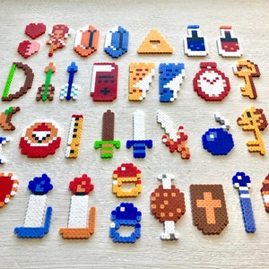 Puede incluir: Una colecci&oacute;n de dise&ntilde;os de pixel art, que incluyen corazones, escudos, espadas, llaves y otros iconos tem&aacute;ticos de juegos. Los art&iacute;culos est&aacute;n hechos de cuentas de pl&aacute;stico de colores, dispuestas sobre una superficie clara.