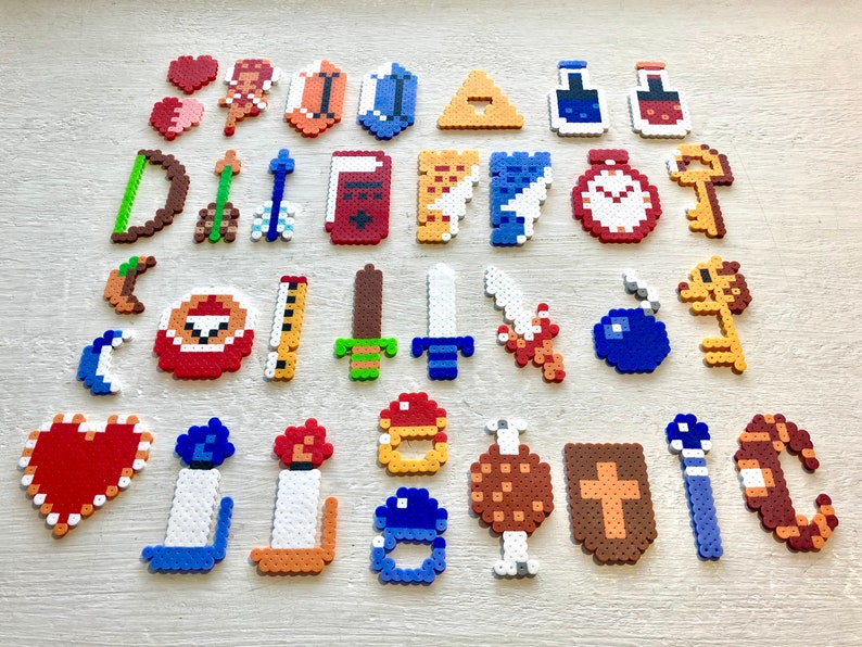 Puede incluir: Una colecci&oacute;n de dise&ntilde;os de pixel art, que incluyen corazones, escudos, espadas, llaves y otros iconos tem&aacute;ticos de juegos. Los art&iacute;culos est&aacute;n hechos de cuentas de pl&aacute;stico de colores, dispuestas sobre una superficie clara.