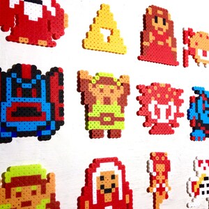May include: A collection of colorful pixel art characters, including a red monster, a yellow triangle, and various figures from a video game. The art is made from small, fused plastic beads, creating a retro aesthetic.