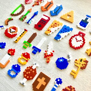 Puede incluir: Una colecci&oacute;n de objetos de juego con tem&aacute;tica de pixel art, que incluye espadas, llaves, pociones y bombas. Los objetos est&aacute;n hechos de coloridas cuentas de pl&aacute;stico y dispuestos sobre una superficie de madera clara. Los objetos son de varios colores, incluyendo rojo, azul, amarillo y verde.