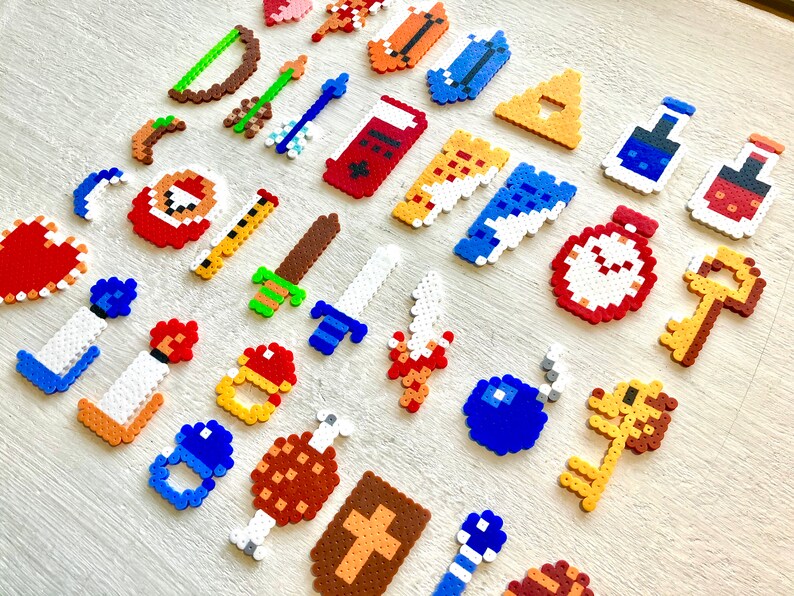 Puede incluir: Una colecci&oacute;n de objetos de juego con tem&aacute;tica de pixel art, que incluye espadas, llaves, pociones y bombas. Los objetos est&aacute;n hechos de coloridas cuentas de pl&aacute;stico y dispuestos sobre una superficie de madera clara. Los objetos son de varios colores, incluyendo rojo, azul, amarillo y verde.