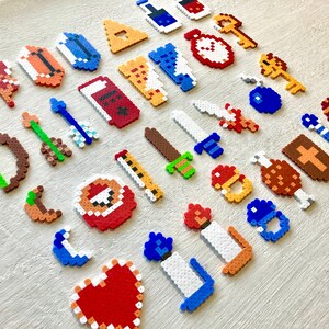 Puede incluir: Una colecci&oacute;n de creaciones de pixel art, que incluyen corazones, espadas, llaves y otros elementos tem&aacute;ticos de juegos. Los art&iacute;culos est&aacute;n hechos de cuentas de pl&aacute;stico de colores, dispuestas sobre una superficie clara.