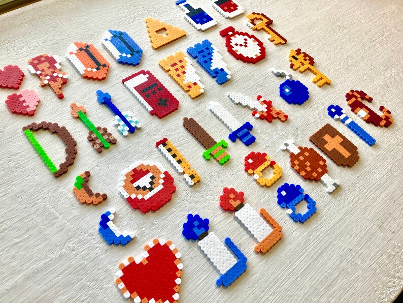 Puede incluir: Una colecci&oacute;n de creaciones de pixel art, que incluyen corazones, espadas, llaves y otros elementos tem&aacute;ticos de juegos. Los art&iacute;culos est&aacute;n hechos de cuentas de pl&aacute;stico de colores, dispuestas sobre una superficie clara.