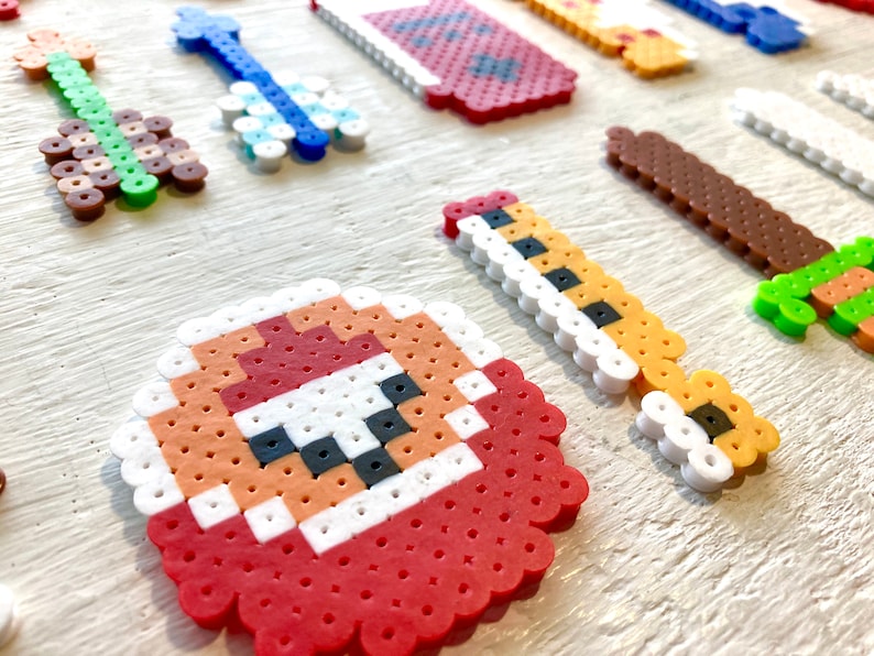 Puede incluir: Una colecci&oacute;n de creaciones de arte pixelado coloridas, que incluyen una guitarra, una llave y una espada, hechas de peque&ntilde;as cuentas de pl&aacute;stico. Los dise&ntilde;os presentan varios colores como verde, azul, marr&oacute;n y rojo, dispuestos sobre una superficie clara.