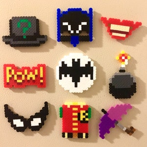 Può includere: Una collezione di calamite pixel art a tema fumetti e supereroi. Include un cappello dell'Enigmista, il simbolo di Batman, il costume di Robin, una nuvoletta "Pow!" e altri elementi iconici dai colori vivaci.