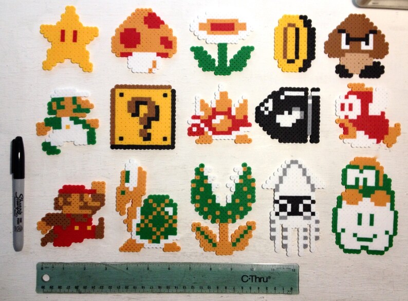Puede incluir: Una colecci&oacute;n de personajes y objetos coloridos de pixel art de la serie de videojuegos Super Mario. Incluye a Mario, Luigi, una estrella, una seta, una moneda, un bloque de interrogaci&oacute;n y otros elementos ic&oacute;nicos. Se incluye un rotulador negro y una regla.