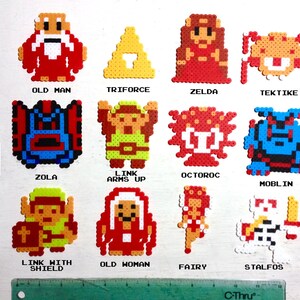 May include: A collection of colorful pixel art characters from the Legend of Zelda video game series. The characters include Old Man, Zelda, Link, and others, all created with small, fused plastic beads. A black marker and ruler are included for scale.
