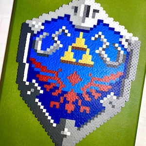 以下が含まれることがあります： 青、赤、黄、グレー、白のピクセル化されたシールドデザインで、ビデオゲームを彷彿とさせます。シールドは緑色の表面に取り付けられています。シールドは中央のエンブレムと詳細なボーダーを備えています。