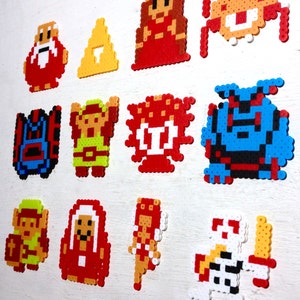 May include: A collection of colorful pixel art characters and symbols, likely from a video game. The designs are made from small, fused plastic beads. The characters include a wizard, a princess, and a hero with a sword.