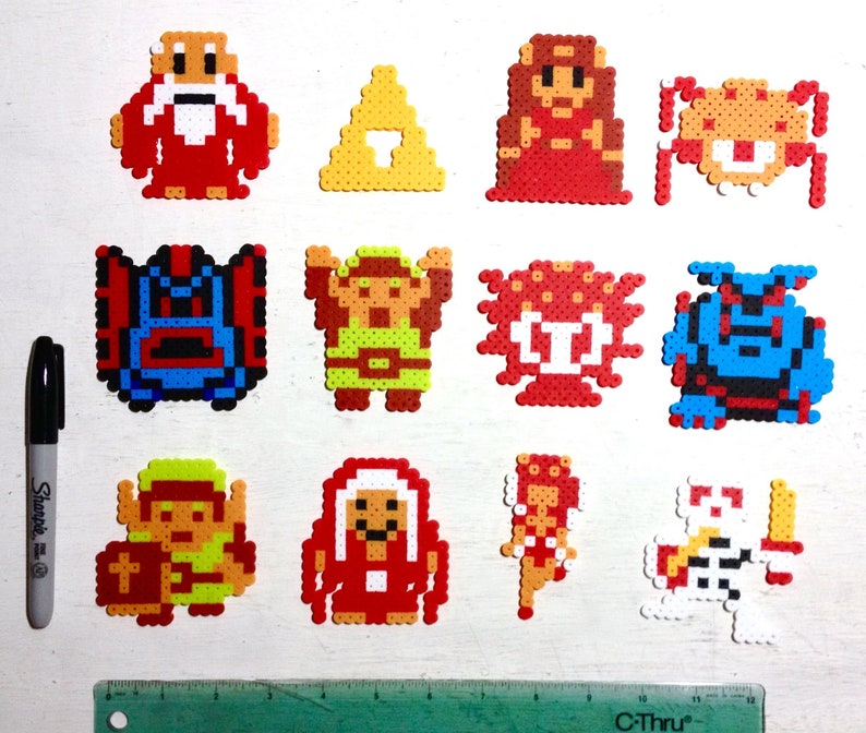 May include: A collection of colorful pixel art characters and symbols, likely from a video game. The figures are made from small, fused beads in various colors, including red, yellow, blue, and white. A black marker and ruler are included for scale.