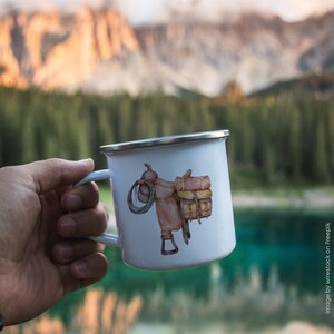 May include: White enamel mug featuring a watercolor illustration of a cowboy saddle with a rope and saddlebags. The mug has a silver rim and a light blue handle. The design is set against a blurred outdoor background.
