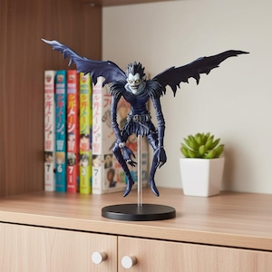 May include: A detailed figurine of a character with dark blue wings and attire, perched on a black base. The figure has a skeletal face with yellow eyes and spiky black hair. The background includes a shelf with manga books and a small potted plant.