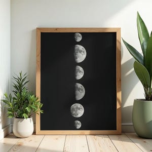 Puede incluir: Una impresión enmarcada que muestra las fases de la luna sobre un fondo negro. Las lunas se representan en diferentes etapas, desde la creciente hasta la llena, en tonos grises y blancos. El marco es de madera clara y la obra de arte se exhibe sobre un suelo de madera.