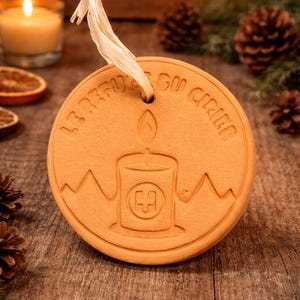 Op de afbeelding: Een ronde, terracotta-kleurige ornament met een kaarsontwerp en de tekst "LE REFUGE DU CHERIE" in reliëf langs de rand. Het heeft een gat aan de bovenkant met een crèmekleurig lint om op te hangen. De achtergrond bevat kaarsen en dennenappels.