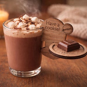 Op de afbeelding: Een dampend glas warme chocolademelk met marshmallows en chocoladeschilfers. Een houten koekjeshouder met de tekst "Have a cookie!" en een stuk chocolade staat naast het drankje. Een brandende kaars staat op de achtergrond.