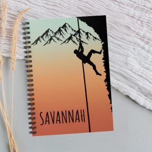 Puede incluir: Cuaderno de espiral con diseño de escalada. La portada presenta la silueta de un escalador en una cuerda sobre un fondo degradado naranja, amarillo y verde. El nombre "Savannah" está impreso en negro.