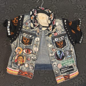 May include: A light-wash denim vest adorned with numerous horror-themed patches, pins, and studs. The collar features a black fabric with orange skulls and white pearls. Patches include text reading Soylent Green is PEOPLE and INVASION OF THE BODY SNATCHERS.