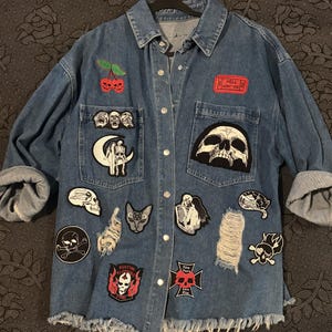 Könnte beinhalten: Ein blaues Jeanshemd mit Knopfleiste, ausgefranstem Saum, Distressed-Stellen und verschiedenen gestickten Aufnähern, darunter Totenköpfe, eine Mondsichel, eine Katze und ein rotes Ticket mit dem Text HELL ADMIT ONE. Langärmlig mit umgeschlagenen Bündchen.