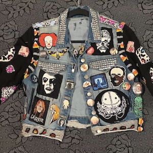 May include: A blue denim jacket with black sleeves featuring various horror-themed patches, metal studs on the collar and chest pockets, and numerous circular pinback buttons. The patches include images of Pennywise, Carrie, and other icons.