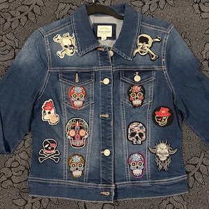 Könnte beinhalten: Eine blaue Jeansjacke mit Knopfleiste, langen Ärmeln und Kragen. Die Jacke ist mit verschiedenen gestickten Aufnähern verziert, darunter mehrere bunte Totenköpfe im Zucker-Stil, klassische Totenköpfe mit gekreuzten Knochen und ein gehörntes Totenkopfmotiv.